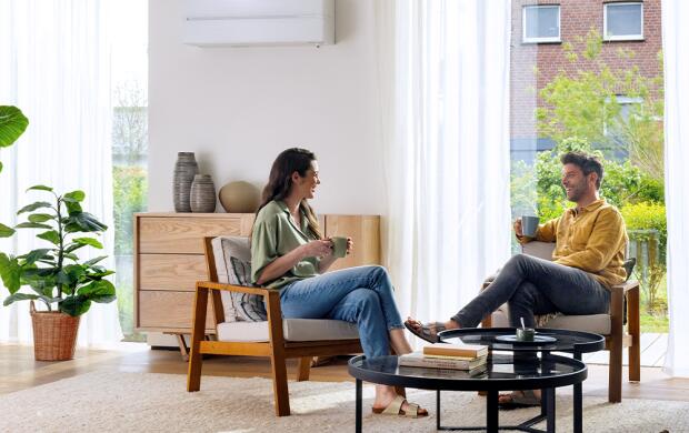 Twee mensen zitten in een lichte woonkamer met een plant, airconditioning en een salontafel.