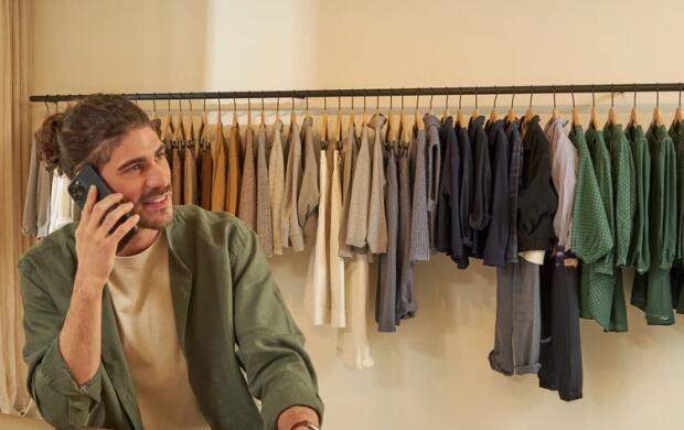 Man telefoneert in kledingwinkel, met hangende kleren en opgevouwen kledingstuk op tafel.