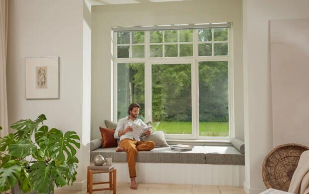 Een man leest een krant op een zitje bij een groot raam met uitzicht op de tuin.