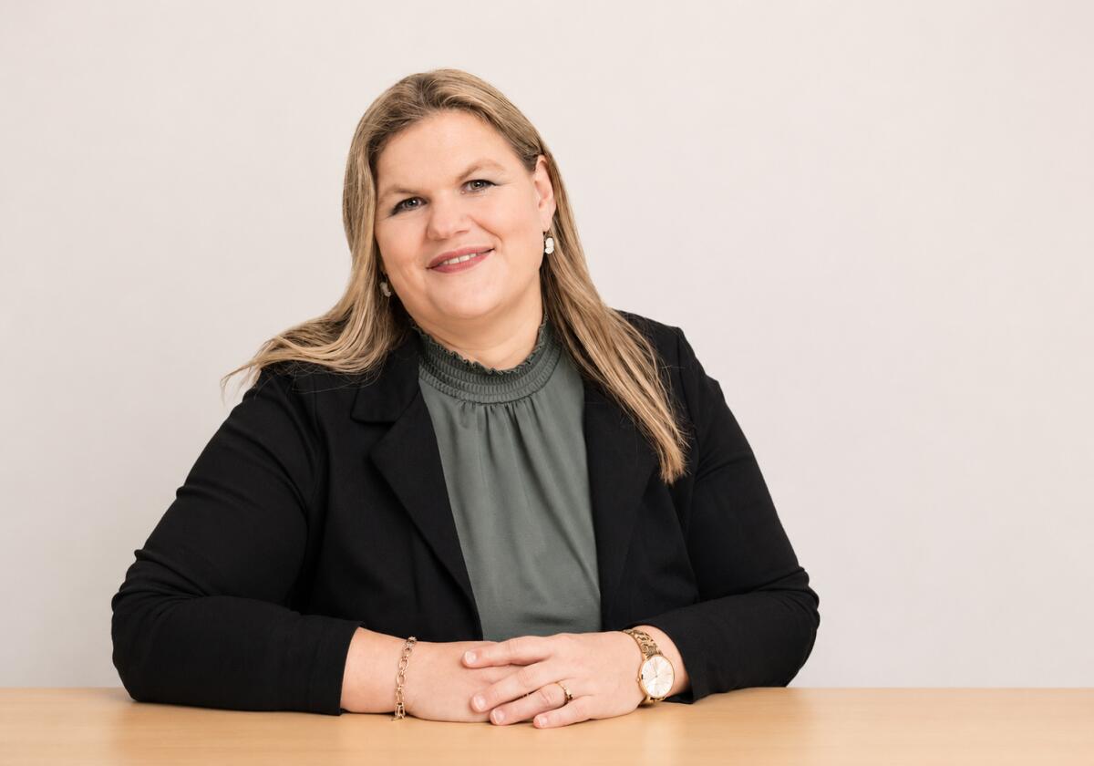 Een lachende vrouw zit aan een tafel, gekleed in een blazer en groene blouse.