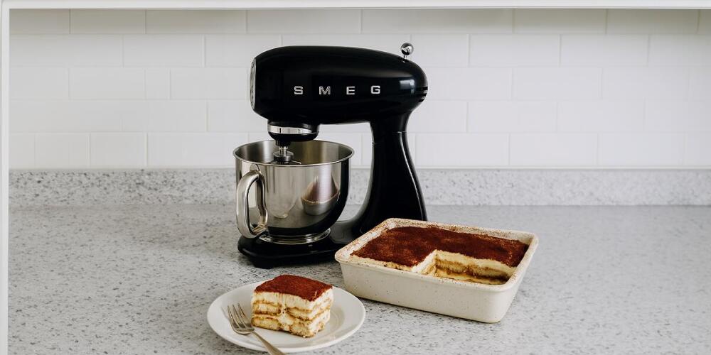 Een keukenmixer naast een schaal met tiramisu en een bordje met een stuk tiramisu.