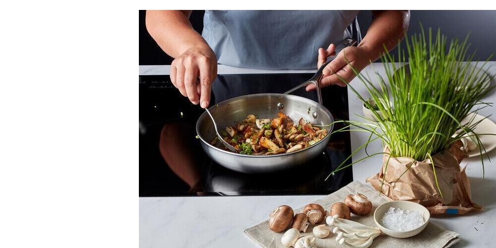 Iemand kookt groenten in een pan op een kookplaat met kruiden en champignons op de voorgrond.