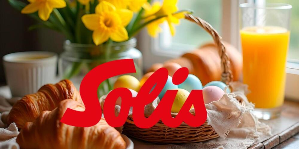 Croissants, gekleurde eieren, bloemen, glas sinaasappelsap en het woord "Solis" bovenop.