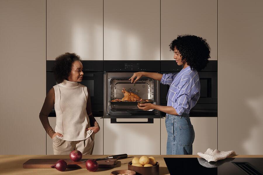 Twee personen in een keuken, een van hen haalt gebraden kip uit een open oven.