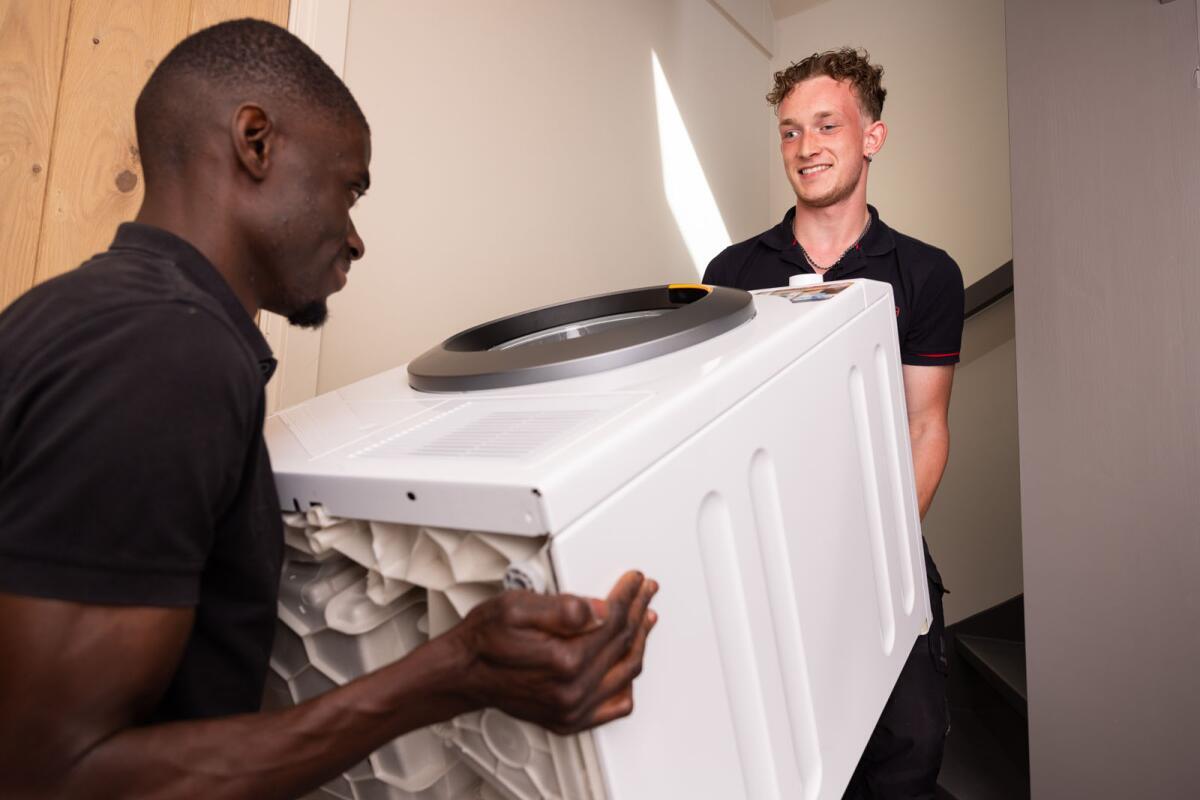 Twee personen tillen samen een wit apparaat, mogelijk een wasmachine, in een gang of trapopgang.