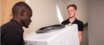 Twee personen tillen samen een wit apparaat, mogelijk een wasmachine, in een gang of trapopgang.