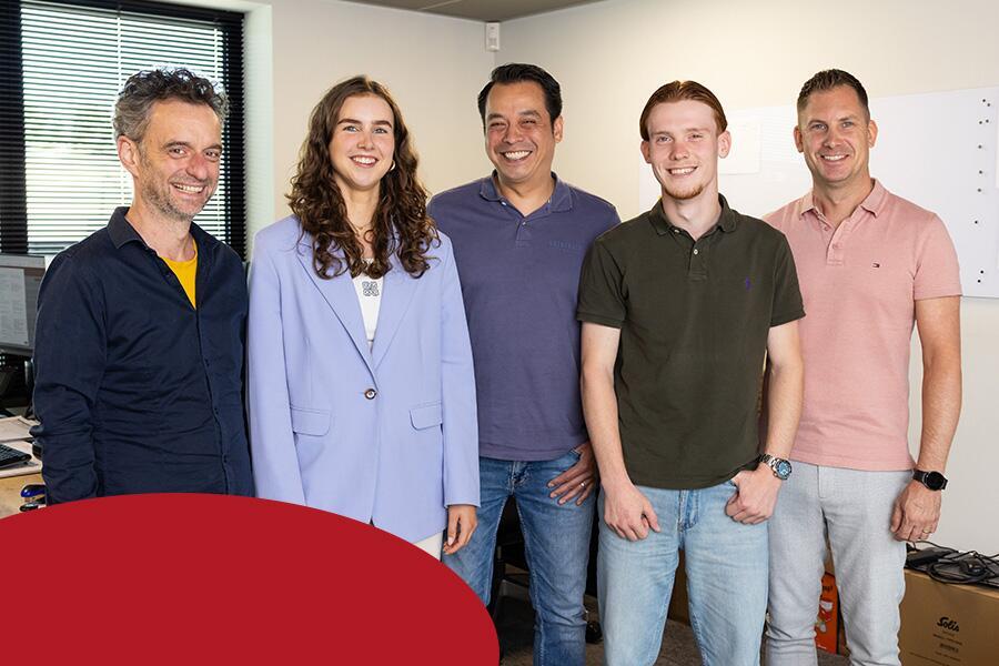 Een groep van vijf mensen staat glimlachend voor de camera in een kantooromgeving.