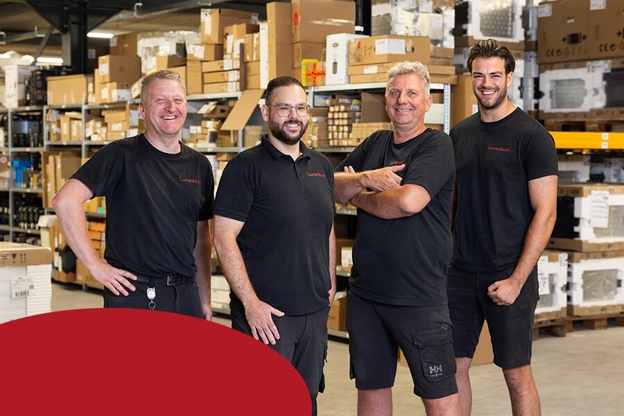 Vier lachende mannen in een magazijn met verschillende dozen op de achtergrond.