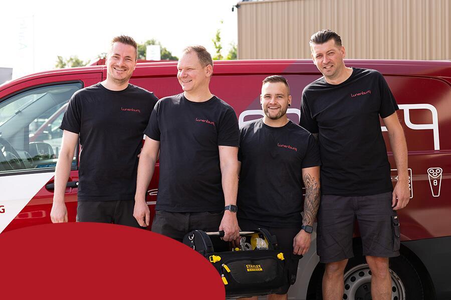 Vier mannen in zwarte shirts poseren lachend voor een rode bestelwagen. Eén houdt gereedschap vast.