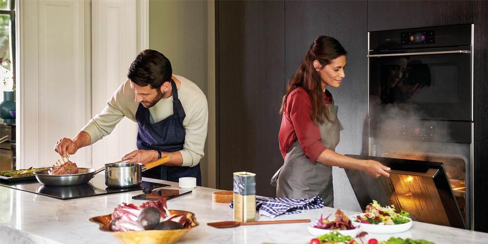 Twee mensen koken in een moderne keuken; een man roert in een pan, een vrouw opent de oven.