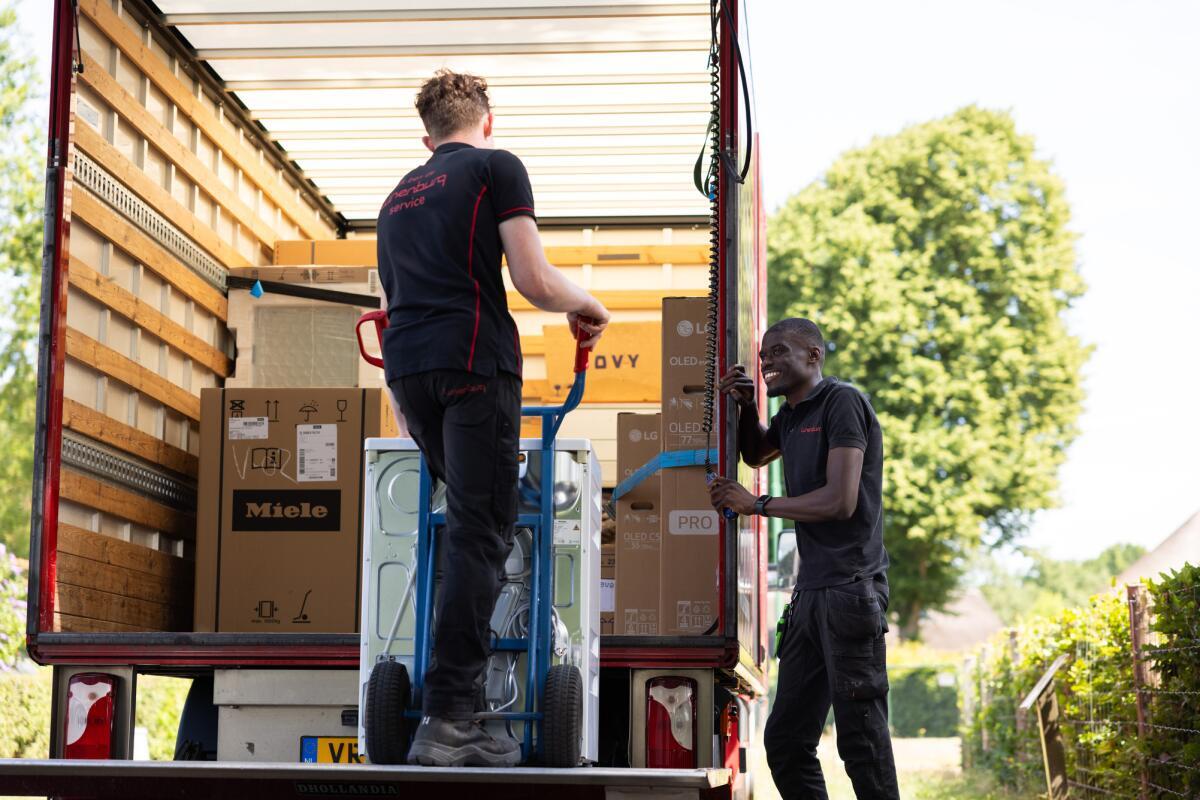 Twee personen laden dozen in een vrachtwagen met een steekwagen op een zonnige dag.