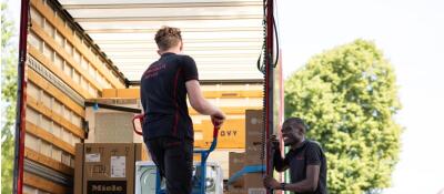 Twee personen laden dozen in een vrachtwagen met een steekwagen op een zonnige dag.