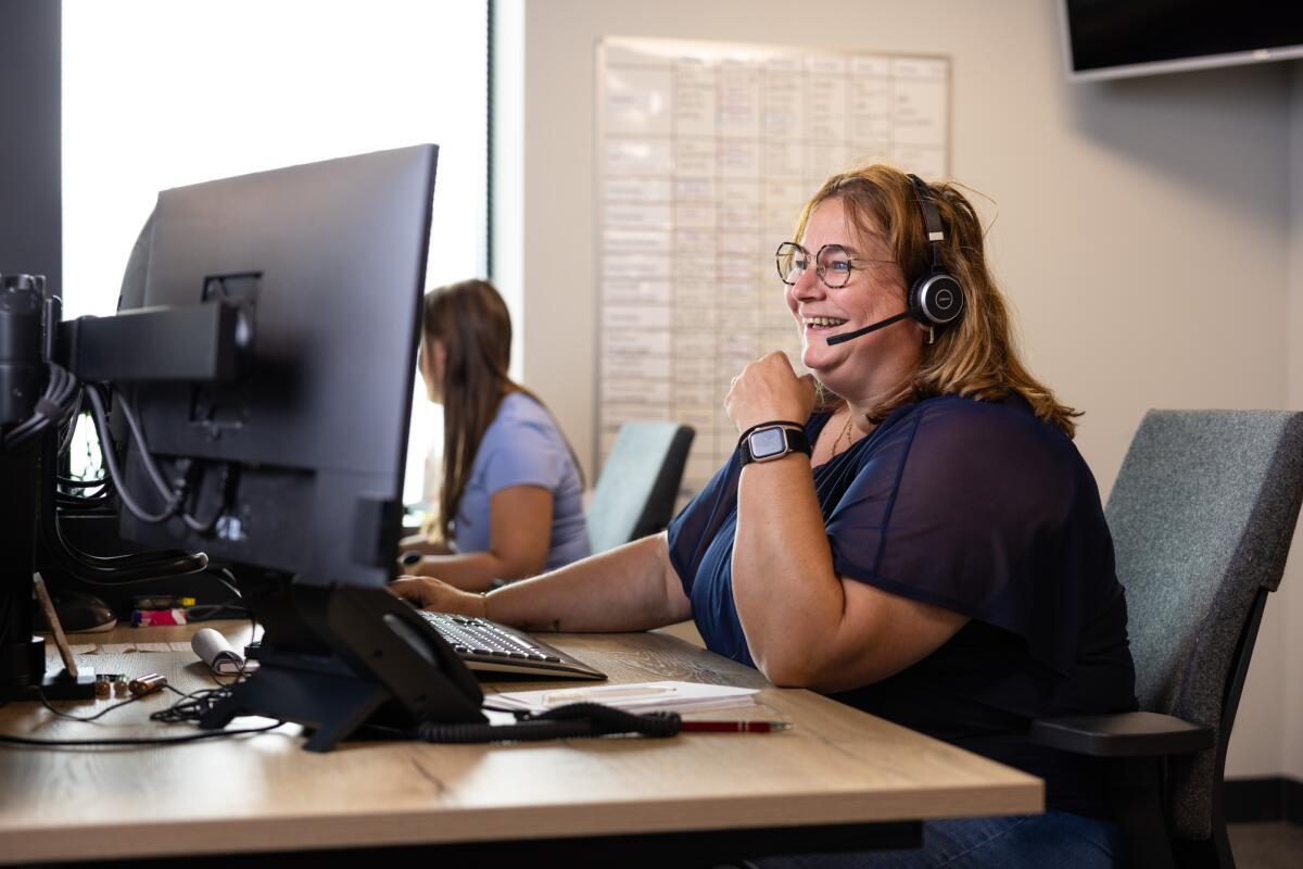 Twee vrouwen werken op kantoor, één draagt een headset en kijkt lachend naar een computerscherm.