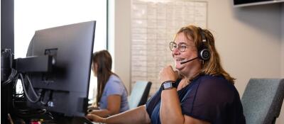 Twee vrouwen werken op kantoor, één draagt een headset en kijkt lachend naar een computerscherm.
